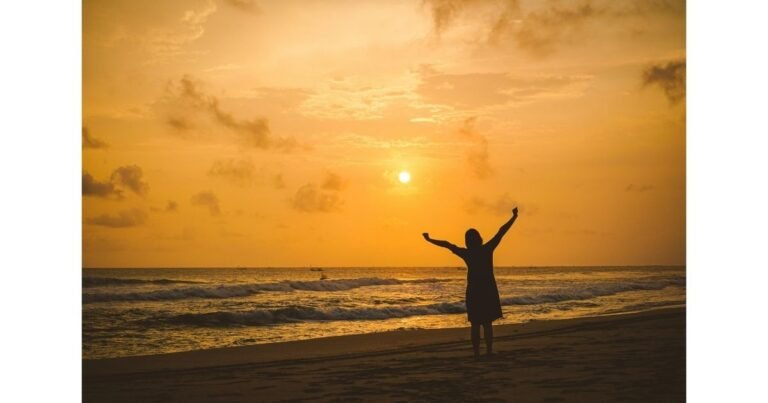 A silhouette of a woman standing on the beach at sunset with her arms raised, symbolizing gratitude and a Christ-centered life.
